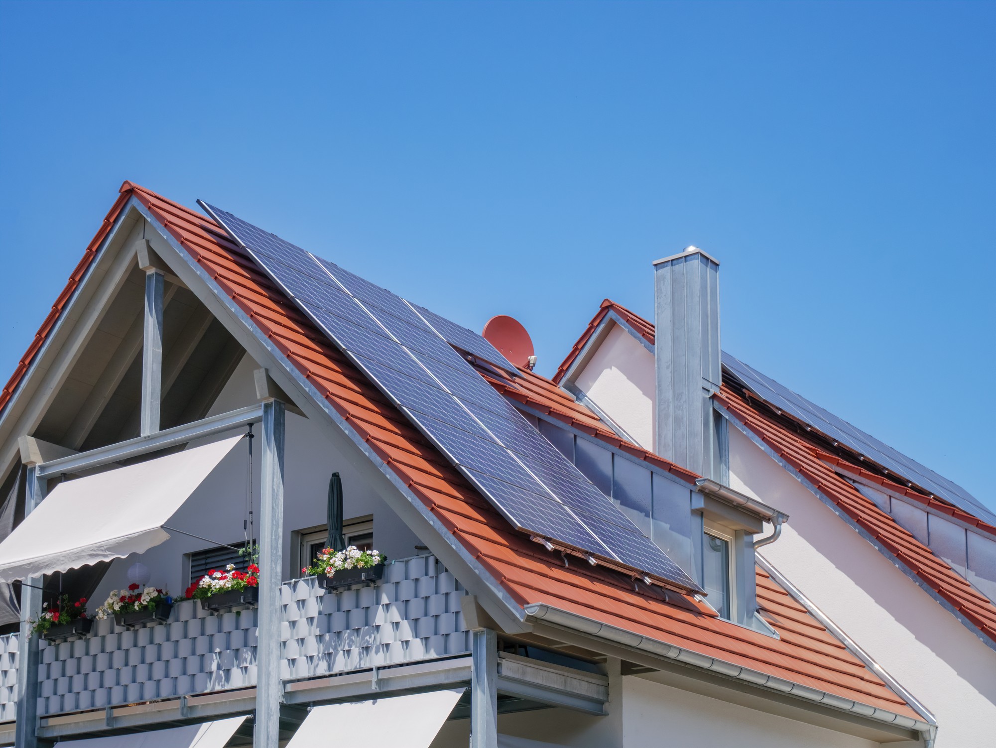 This is an image of the side of a building, which appears to be a residential house, with a focus on the roof area. The roof is equipped with a series of solar panels, which are used to convert sunlight into electricity as part of a renewable energy system for the home. The roof tiles are terracotta in colour, contrasting with the dark blue of the solar panels.

The house also features a white balcony with a railing adorned with flower boxes containing red and white flowers, adding a decorative and homey touch. Additionally, there's a retractable sunshade extended over the balcony area, providing shade for the space below. A chimney stack and a red satellite dish are also visible on the roof. The sky is clear and blue, indicating a sunny day which is ideal for generating solar power.