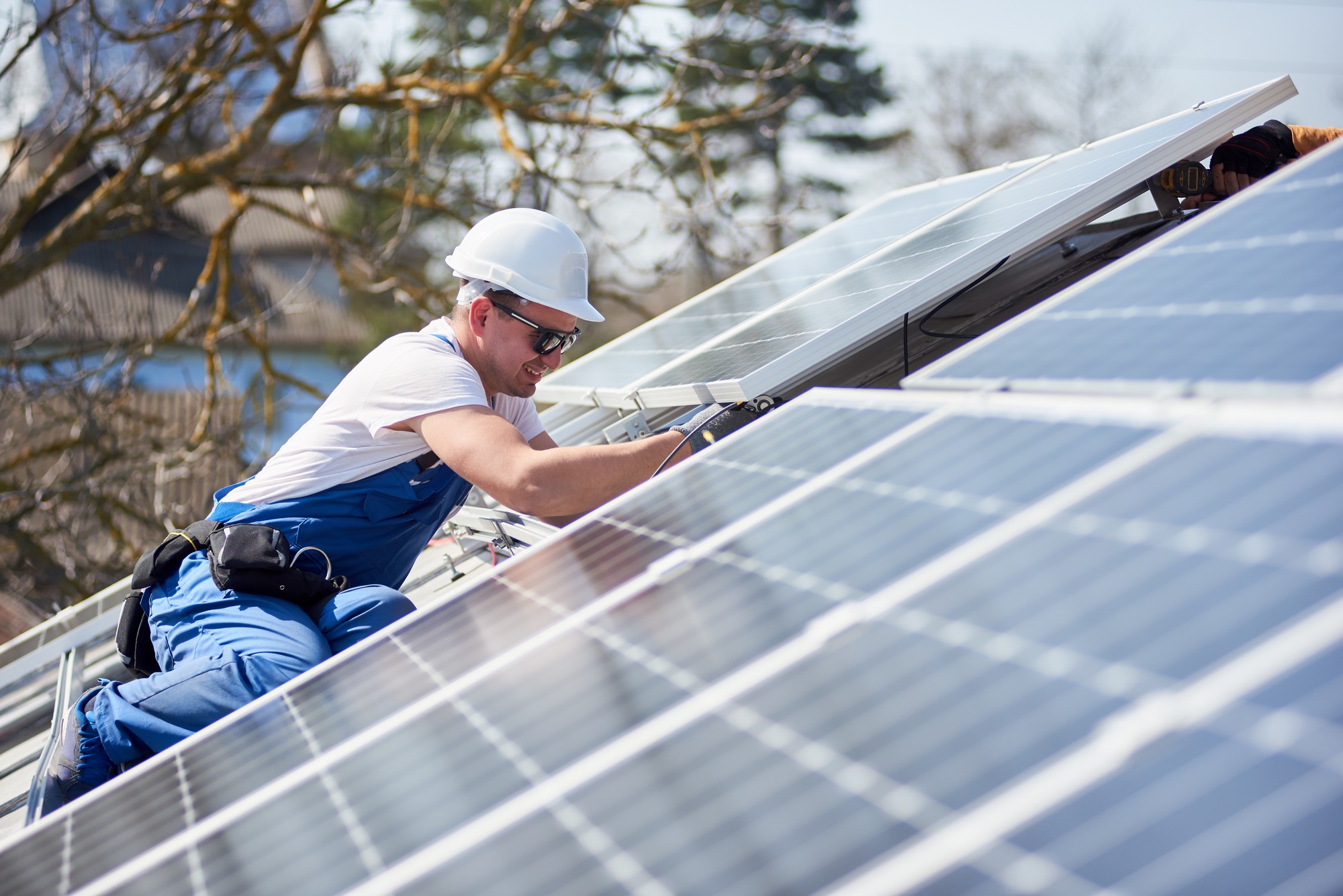 The image shows a worker wearing a white safety helmet, sunglasses, and blue work gear installing or maintaining solar panels on a rooftop. The worker is using a tool, presumably for securing the panels in place, and appears to be focused on the task at hand. The day is sunny, which is conducive to activities involving solar panels, as they depend on sunlight to generate electricity. Trees and a clear sky can also be seen in the background, suggesting a suburban setting.