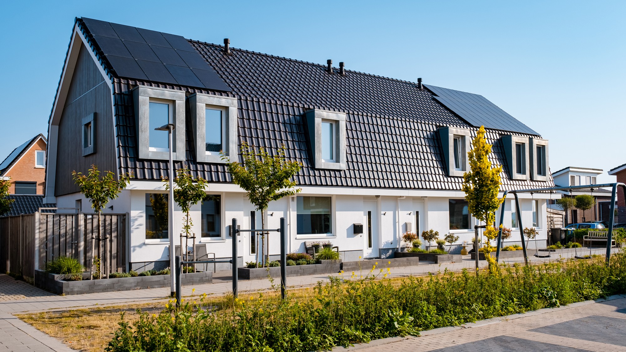 The image shows a row of modern semi-detached houses. The houses feature dark pitched roofs, some of which are equipped with solar panels, suggesting a focus on sustainability and energy efficiency. The facades are white with wooden details, and each house has large windows. There's a clear sky, indicating a bright, sunny day. The front gardens are well-kept, with small plants and seating areas, and each property is delineated by fencing or hedging. In front of the houses, there is a sidewalk and a bike rack, implying that this is a bike-friendly area. Additionally, young trees are planted along the sidewalk, which may provide shade and aesthetic appeal as they mature. The environment appears to be a tranquil residential neighborhood.