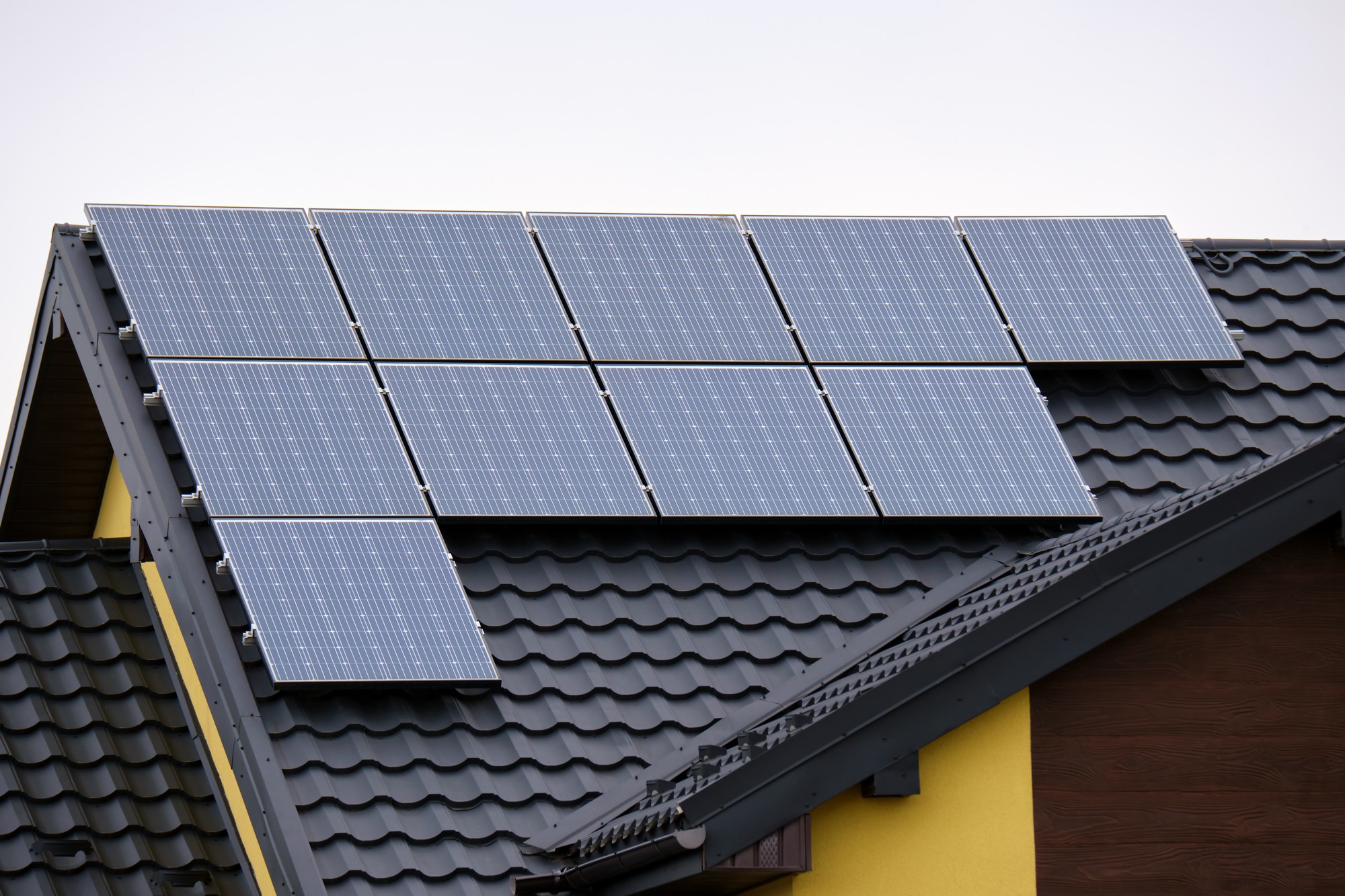 The image shows a set of solar panels installed on the roof of a house. The panels are arranged in a tidy grid pattern and appear to be photovoltaic panels, which are used to convert sunlight into electrical energy. The roof has dark-coloured tiles, and the bottom edge of the roof has a gutter. One side of the house has a partial view of a wall with a yellow façade and a section covered in brown material, possibly wooden siding or panels. The day seems overcast, as the sky is gray, suggesting diffused lighting conditions.