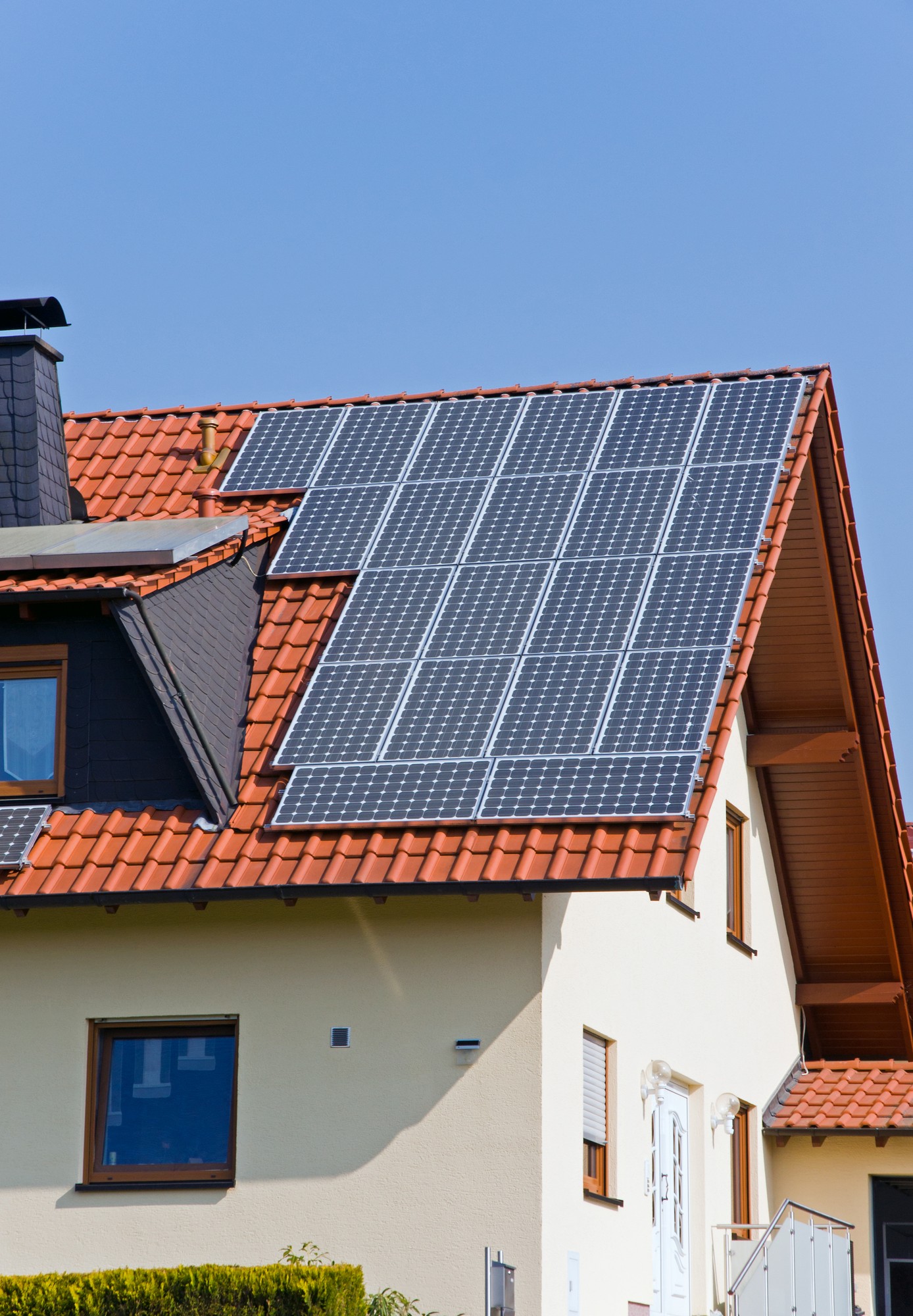 In this image, you can see a part of a house with a pitched roof that has solar panels installed on it. The roof tiles are terracotta in colour, and the house is painted in a light beige with white trim around the windows and on the railing of the entrance stairway. The sky is clear, suggesting a sunny day which is ideal for solar energy production. The presence of solar panels indicates a commitment to renewable energy sources for the household's electricity needs.