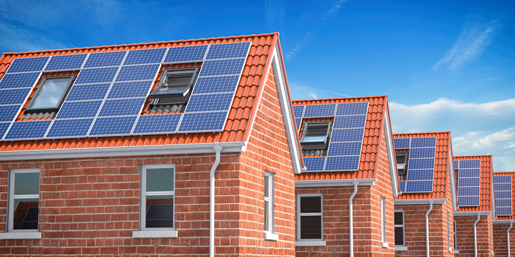 This image shows a row of modern brick houses with orange-red tiled roofs, each fitted with solar panels. The houses have several windows and roof windows, and the image suggests that these houses may be equipped with technology for sustainable energy production, indicative of a focus on renewable energy and energy efficiency in residential construction. The sky is blue with a few clouds, indicating a clear day which is ideal for solar power generation.