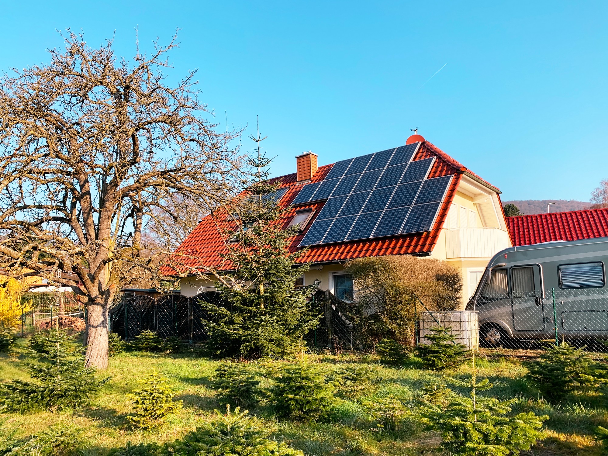 This image features a residential setting with a clear blue sky. There's a house with a red-tile roof equipped with solar panels, indicating a use of renewable energy. In front of the house is a garden with various plants and trees, including an apparent leafless tree to the left, which suggests it might be either autumn or winter, or perhaps an early spring before the foliage has returned. Also visible is a caravan parked to the right side of the house, perhaps used for travel or as a mobile home extension. The overall ambiance of the image is sunny and peaceful.